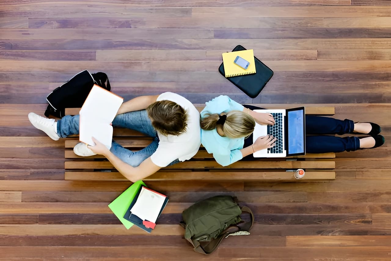 Two students sitting on a bench with books and laptop